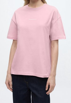 Personne portant un t-shirt ample rose clair à manches courtes avec un petit texte blanc sur la poitrine et un jean bleu foncé, debout devant un fond uni.