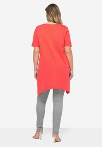 Túnica de coral con mangas cortas, dobladillo redondeado y un corte relajado, combinada con jeans ajustados grises y sandalias de tacón plateadas.