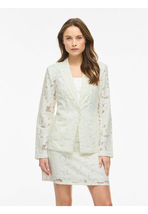 Femme aux longs cheveux bruns portant un blazer blanc texturé et une jupe assortie, debout sur un fond blanc uni.