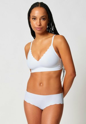 Woman with long braided hair wearing matching white scalloped-edge bra and underwear against plain background.