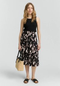 Juodas be rankovių viršus, raštuotas ilgas plisė sijonas juodų ir rožinių atspalvių, laikoma smėlio spalvos pirkinių krepšys ir dėvimos juodos sandalės.