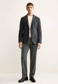 Blazer a spina di pesce grigio su un maglione crema, abbinato a pantaloni grigi e mocassini neri. Design su misura con una silhouette strutturata.