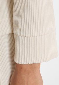 Beige gerippter Stoff, der eine Nahaufnahme eines Ärmelsaum mit horizontalen Linien zeigt, glatte Textur und abgerundete Kante.