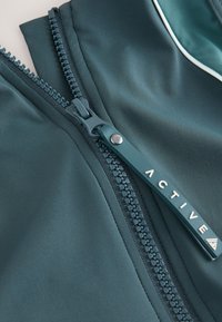 Gros plan sur une fermeture éclair vert foncé avec un curseur étiqueté "ACTIVE" sur un tissu assorti avec un passepoil blanc.