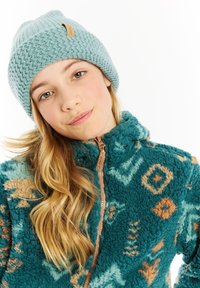 Giacca in pile teal con motivi geometrici, chiusura frontale con zip e colletto alto; abbinata a un berretto di lana azzurro chiaro con etichetta.