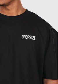 Fekete pamut póló, crew nyakkivágással és egyszerű fehér "DROPSIZE" logóval a mellkason, normál fazonban.