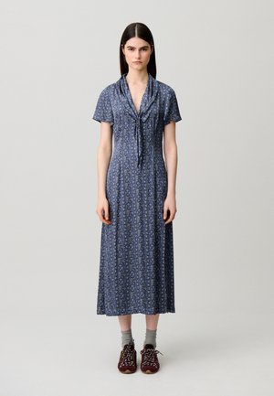 Jeune femme aux cheveux lisses et foncés porte une robe midi bleue à motifs avec des manches courtes, des chaussettes grises et des baskets bordeaux, debout immobile.