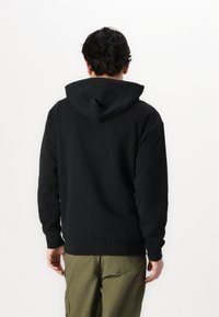Czarny sweatshirt z kapturem, z długimi rękawami, ściągaczami przy mankietach i u dołu. Nosi się go na oliwkowe spodnie, ukazując gładką teksturę tkaniny.
