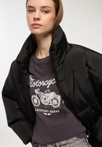 Giacca a vento nera con colletto alto, caratterizzata da una texture liscia. Sotto, una t-shirt grafica scura mostra un design di moto in bianco.