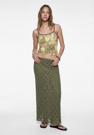 Kobieta ubrana w żółty top w kwiaty, długą oliwkowozieloną koronkową spódnicę, czarne klapki i bransoletki, stojąca na tle jednolitego białego tła.