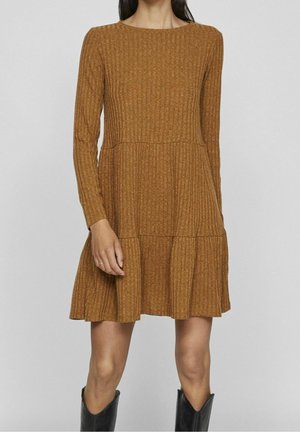 Femme portant une robe en tricot marron à manches longues et côtelée, avec une jupe à volants, associée à des bottes noires mi-mollet.