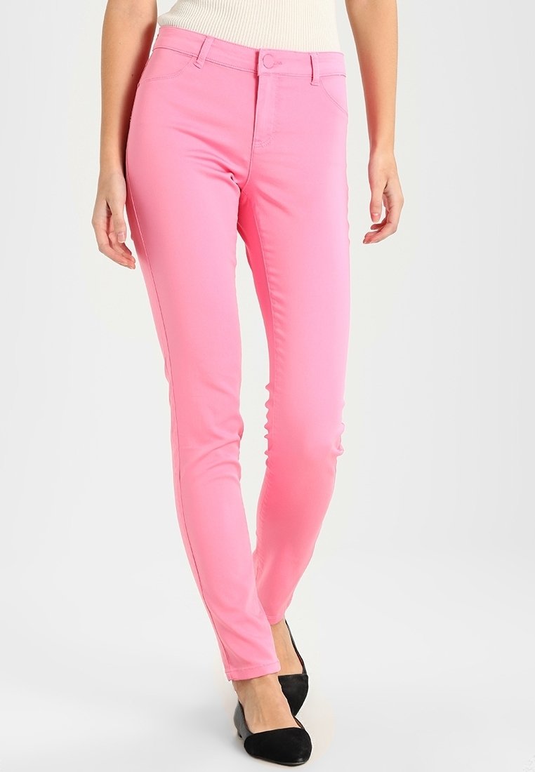 Jean skinny rose à texture lisse, avec une fermeture boutonnée et à glissière, passants pour ceinture, et une coupe élégante, porté avec des chaussures noires.