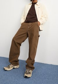 Pantalon marron à jambes larges avec poches latérales, associé à une veste crème boutonnée et une chemise marron foncé. Baskets athlétiques beige et gris.