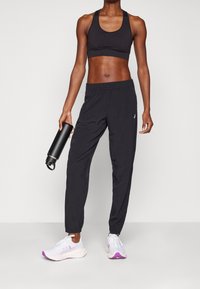 Svart sport-bh i atletisk stil kombinerad med svarta tapered joggerbyxor. Modell som håller en svart vattenflaska; bär vita sneakers med lila detaljer.