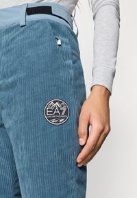 Calças de veludo azul com uma superfície texturizada, apresentando um patch de logo circular em preto e branco na coxa e uma cintura azul clara.