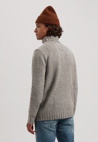 Grijze, gebreide turtleneck trui met ribbelige onderkant en kraag, gecombineerd met blauwe spijkerbroek. Een bruine beanie wordt op het hoofd gedragen.
