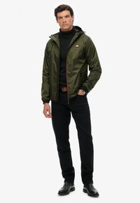 Grön nylonjacket med huva, röda dragkedjedetaljer, bärs över en svart polotröja. Svarta byxor och stövlar fulländar outfiten.