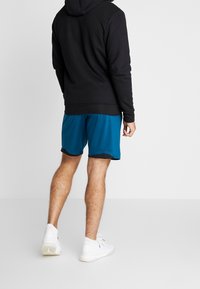 Sudadera con capucha negra, pantalones cortos deportivos color azul verdoso con ribete negro y zapatillas blancas. Ropa deportiva casual vista desde la parte trasera.