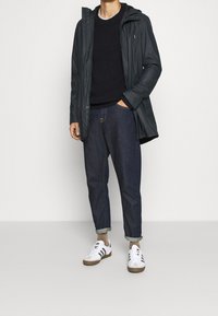Veste à capuche noire, pull côtelé sombre, jeans foncés retroussés et baskets blanches à rayures noires. Superposition simple et fonctionnelle pour un temps plus frais.