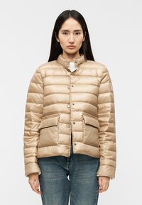 Μπεζ μπουφάν puffer με γιακά που στέκεται, κουμπιά κούμπωσης, δύο μπροστινές τσέπες και οριζόντιο μοτίβο ραφής. Ομαλή, γυαλιστερή υφή υφάσματος.
