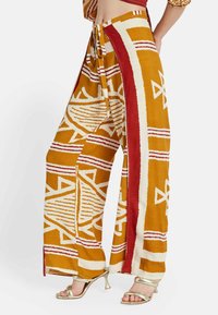 Pantalones de pierna ancha con estampado en color mostaza, con acentos en rojo y blanco. Tejido texturizado con diseños geométricos y aberturas laterales.