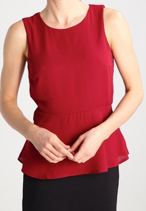 Femme portant un chemisier rouge sans manches avec bas volanté et jupe noire, mains doucement jointes devant le corps.