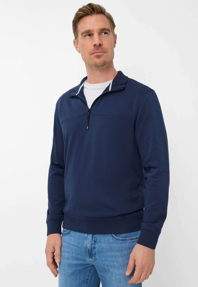Mann trägt einen marineblauen Sweatshirt mit Reißverschlusskragen über einem weißen Hemd und hellblauen Jeans, steht vor einem schlichten weißen Hintergrund.