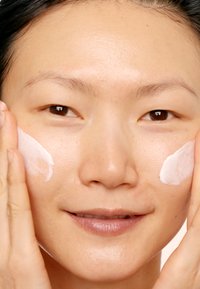 Hvit krem påført kinnet, som avslører en jevn hudtekstur. Lys bakgrunn fremhever ansiktstrekkene og påføringsmetoden.