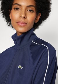Giacca zip-up blu navy con colletto alto, caratterizzata da dettagli in piping bianco e un logo di coccodrillo verde sul lato sinistro. Tessuto liscio.