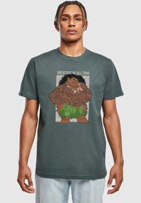 Dark green t-shirt featuring a graphic of a fictional character with intricate tattoos, wearing a leaf skirt, with the text "GREATEST OF ALL TIME."