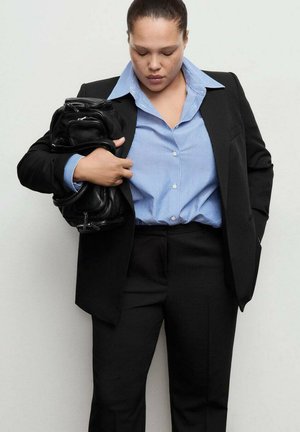 Black tailored suit jacket over a blue and white striped shirt, holding a black textured bag, against a plain light background.
