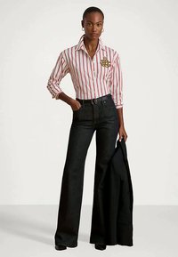 Camisa a rayas blanca y roja con bordado dorado, combinada con pantalones de mezclilla oscuros de tiro alto y acampanados. Chaqueta negra sostenida en la mano izquierda.