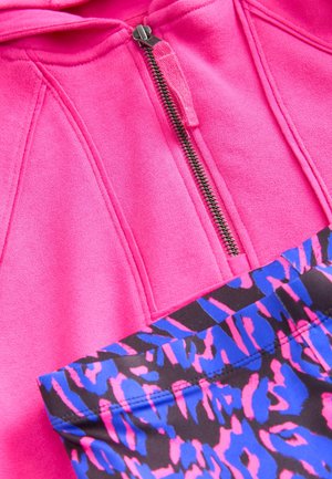 Fuchsiová mikina na zip s kartáčovanou texturou; vzorovaná černá látka s růžovými a modrými pruhy pod ní. Viditelný kovový detail zipu.