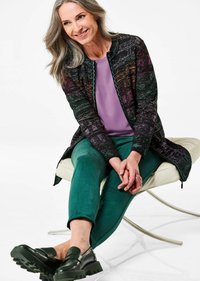 Multikleurige lange cardigan met een textuurpatroon, gecombineerd met een effen paarse top en groene broek. Zwarte instappers met dikke zolen.