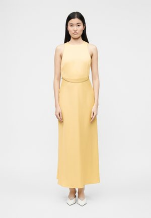 Frau mit langen schwarzen Haaren, die ein ärmelloses, hellgelbes Maxikleid und weiße spitze Schuhe trägt und vor einem schlichten hellen Hintergrund steht.