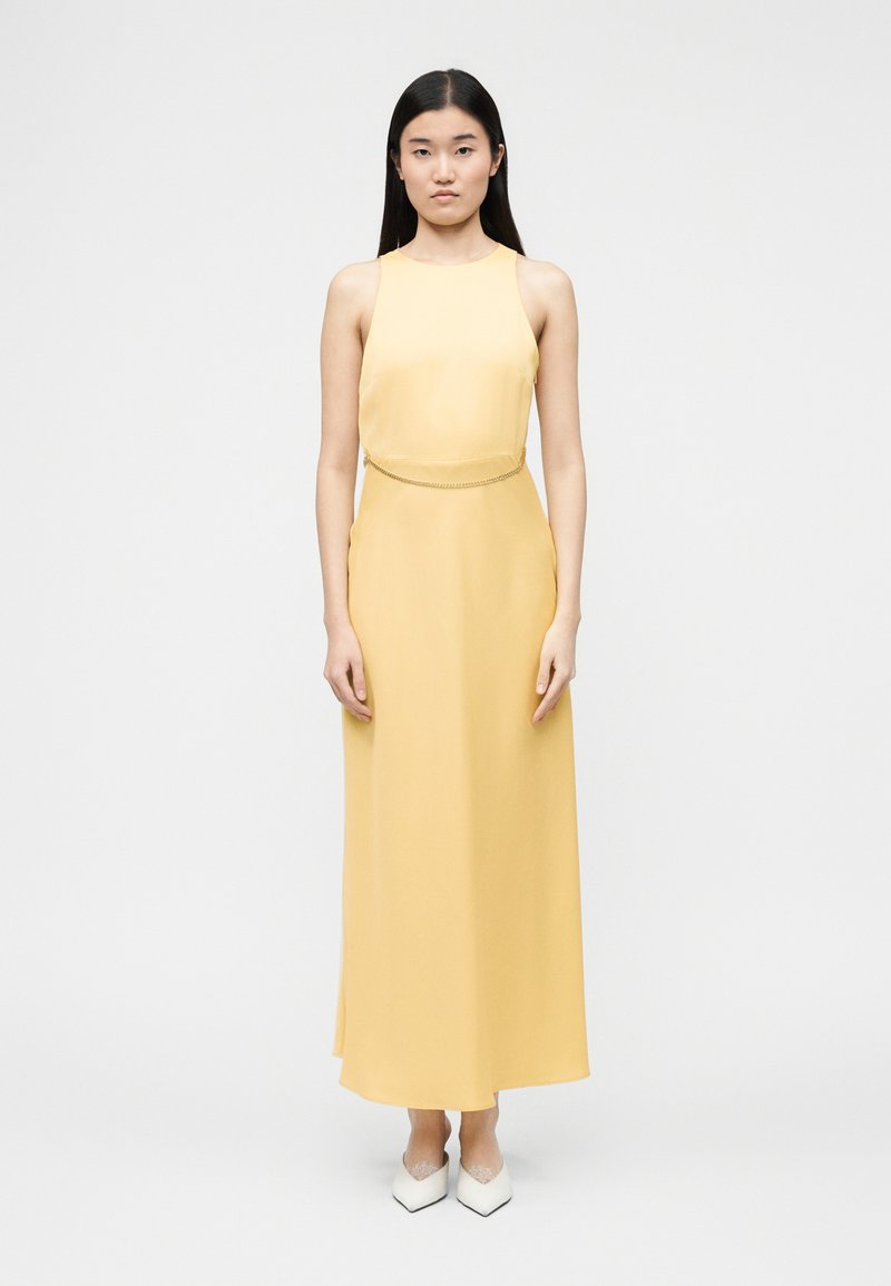 Femme aux longs cheveux noirs portant une robe maxi sans manches jaune pâle et des chaussures blanches à bouts pointus, debout devant un fond clair uni.