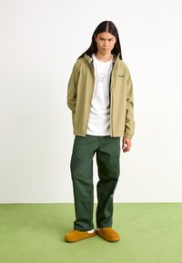 Jachetă impermeabilă de culoare verde oliv, tricou alb cu grafică, pantaloni verzi închis și saboți maroni pe un covor verde deschis. Design simplu și funcțional.