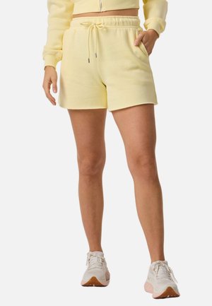 Femme portant un short jaune pâle avec cordon de serrage et un sweat court assorti, baskets blanches, debout devant un fond blanc.