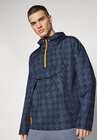 Chaqueta con capucha de color navy, con una tela estampada, que presenta un bolsillo frontal y un zipper naranja, fabricada con un material suave y ligero.