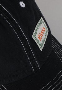Fekete sapka fehér varrással, amelyen egy zöld szegélyű és narancssárga "Rivvia" feliratú téglalap alakú címke található. Pamut anyagból készült, strukturált kialakítással.
