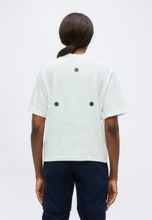 Personne aux cheveux foncés attachés en arrière portant une chemise blanche à manches courtes avec trois points noirs dans le dos et un pantalon sombre, vue de dos.