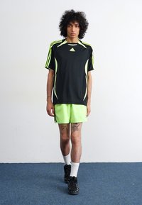 Sort træningsskjorte med limegrønne accenter og striber, parret med limegrønne shorts. Modellen har sorte sneakers og hvide sokker på.