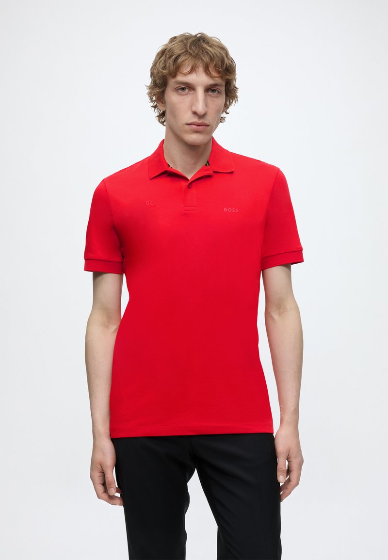 Jeune homme aux cheveux blonds bouclés porte un polo rouge à manches courtes et un pantalon noir, debout devant un fond blanc uni.