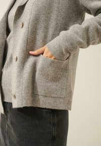 Cárdigan de lana gris con diseño de cuello, dos bolsillos frontales y cierres de botones. La tela parece suave con una textura fina.