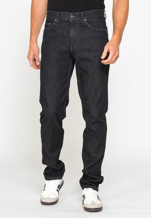 Uomo con jeans neri slim fit e scarpe da ginnastica bianche, in piedi davanti a uno sfondo bianco, mani rilassate ai lati.