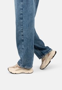 Baskets beiges avec des accents en maille et en cuir, semelles rembourrées et design épais, associées à un jean en denim bleu.