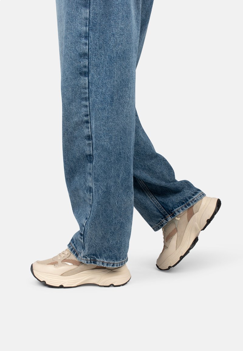 Baskets beiges avec des accents en maille et en cuir, semelles rembourrées et design épais, associées à un jean en denim bleu.