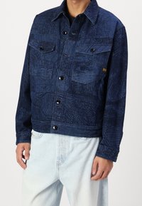 Mörkblå denimjacka med ett texturerat mönster, två bröstfickor och knäppning med knappar. Avslutad med svarta knappar och en kortare skärning.