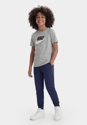 Niño sonriente con cabello rizado, vestido con una camiseta gris de Nike, pantalones deportivos azul marino y zapatillas blancas altas, de pie sobre un fondo blanco liso.