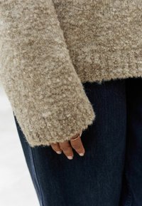 Beige texturerad tröja med lös passform, som har mjukt, fluffigt tyg och rå fåll, bärs över mörk denimjeans, hand med ring synlig.